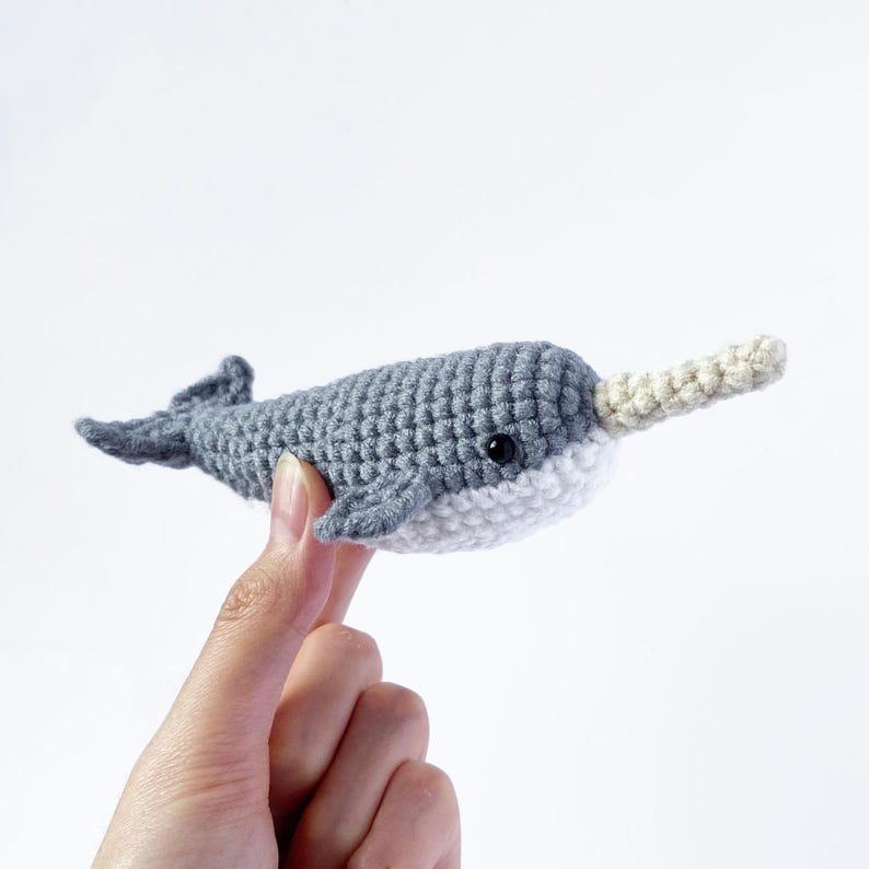 May include: A gray and white crocheted narwhal plush toy with a small black eye. The toy is being held by a hand.