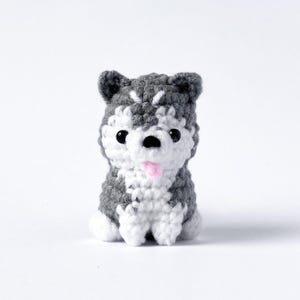Peut inclure: Figurine de chien husky au crochet, en fil gris et blanc. Le chien a des yeux noirs et une langue rose, et est assis. L'objet artisanal est sur fond blanc.