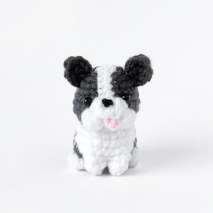 Puede incluir: Un pequeño perro de peluche tejido a crochet con pelo blanco y negro. El perro tiene la lengua rosa que sobresale y ojos negros.