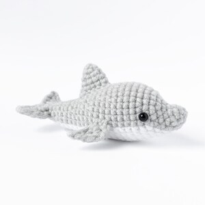 May include: A gray crocheted dolphin plush toy with black eyes. The dolphin is lying on its side with its head turned to the right.