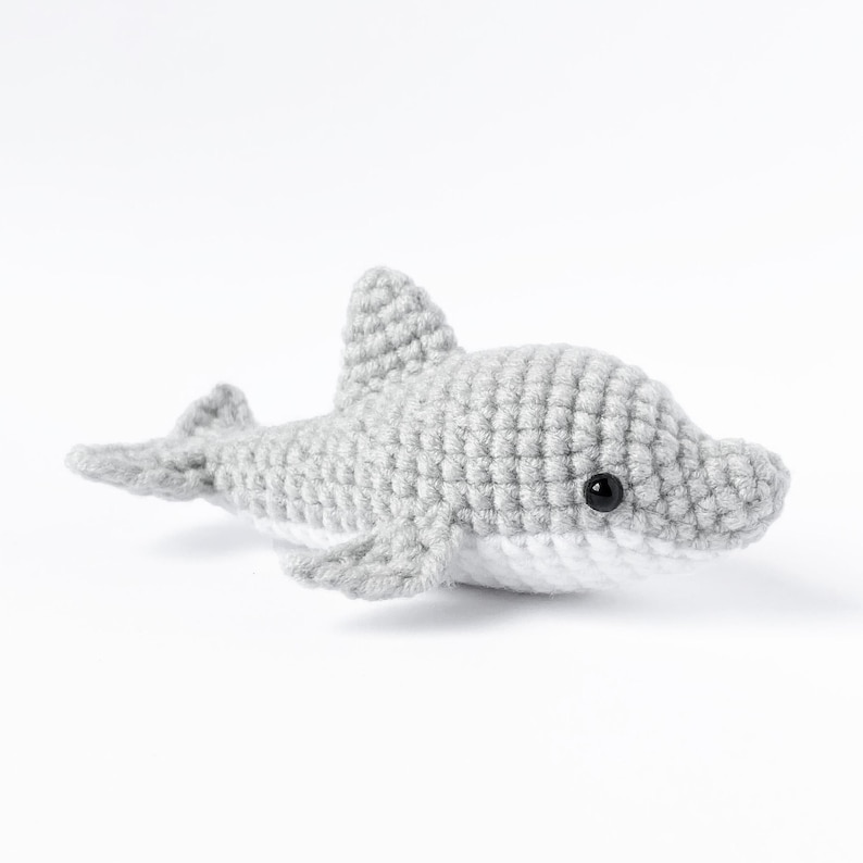 May include: A gray crocheted dolphin plush toy with black eyes. The dolphin is lying on its side with its head turned to the right.