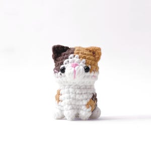 Op de afbeelding: Een klein gehaakt kattenfiguurtje met een lapjeskatpatroon van witte, bruine en beige garenvlekken. De kat heeft twee zwarte kraaloogjes, een klein roze geborduurd neusje en puntige oortjes, gemaakt met een textuursteek.