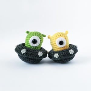 Puede incluir: Dos juguetes extraterrestres de crochet en naves espaciales con forma de platillo volante. Un extraterrestre es verde y el otro es amarillo. Ambos extraterrestres tienen un gran ojo negro.