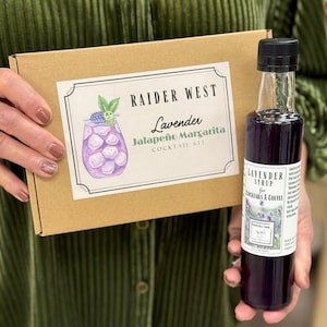 May include: A brown cardboard box labeled Raider West Lavender Jalapeño Margarita Cocktail Kit with a purple drink illustration, paired with a tall clear glass bottle of dark purple lavender syrup featuring a white label with floral imagery.