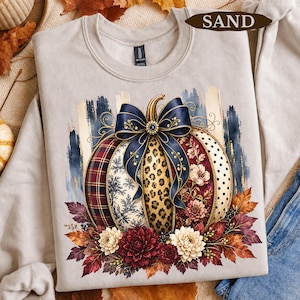 Op de afbeelding: Een zandkleurige sweater met ronde hals en een afbeelding van een pompoen versierd met een grote marineblauwe strik. De pompoensegmenten tonen diverse patronen zoals ruitjes, luipaardprint, bloemen en stippen, omringd door herfstbladeren.