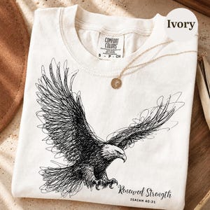 Könnte beinhalten: Ein elfenbeinfarbenes T-Shirt mit einer schwarzen, krakelartigen Grafik eines fliegenden Adlers. Der Text "Renewed Strength" und "ISAIAH 40:31" ist unter dem Adler aufgedruckt. Das T-Shirt hat ein Comfort Colors Etikett und einen goldenen Anhänger.