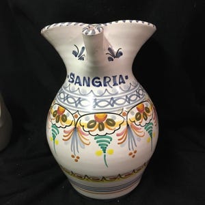 May include: White ceramic jug with a spout, displaying the word "SANGRIÁ" in blue. The jug is decorated with colourful floral and geometric patterns in yellow, orange, green, and blue. A decorative item for serving drinks.