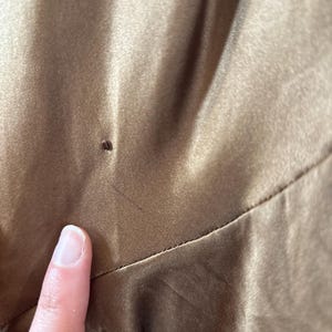 May include: Close-up of a section of a brown satin fabric. The fabric has a smooth, shiny texture with subtle variations in color. A finger is touching the fabric, highlighting its texture and sheen. The image focuses on the details of the material.