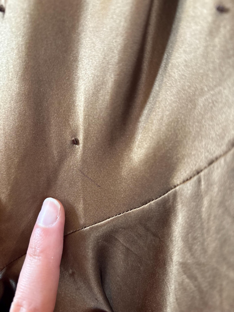 May include: Close-up of a section of a brown satin fabric. The fabric has a smooth, shiny texture with subtle variations in color. A finger is touching the fabric, highlighting its texture and sheen. The image focuses on the details of the material.