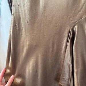 May include: A close-up of a bronze-colored satin dress. The fabric has a subtle sheen and a flowing drape. The dress features a layered design on the side and delicate embellishments near the shoulder. The overall style suggests formal wear.
