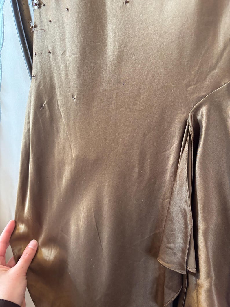 May include: A close-up of a bronze-colored satin dress. The fabric has a subtle sheen and a flowing drape. The dress features a layered design on the side and delicate embellishments near the shoulder. The overall style suggests formal wear.