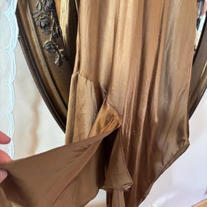 May include: A flowing, bronze-colored dress with a high slit. The dress has a silky, lustrous appearance and a subtle sheen. The fabric drapes elegantly, suggesting a vintage or formal style. The dress is shown against a decorative mirror.