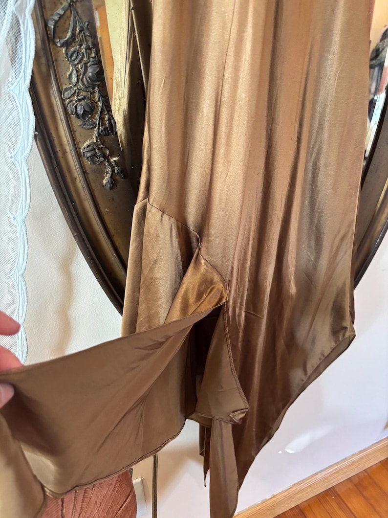 May include: A flowing, bronze-colored dress with a high slit. The dress has a silky, lustrous appearance and a subtle sheen. The fabric drapes elegantly, suggesting a vintage or formal style. The dress is shown against a decorative mirror.