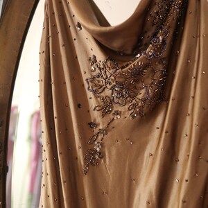 May include: A flowing, champagne-colored dress with a cowl neckline. The dress is adorned with intricate beadwork and sequins in shades of brown and silver, creating a shimmering effect. The fabric appears to be a soft, draping material.