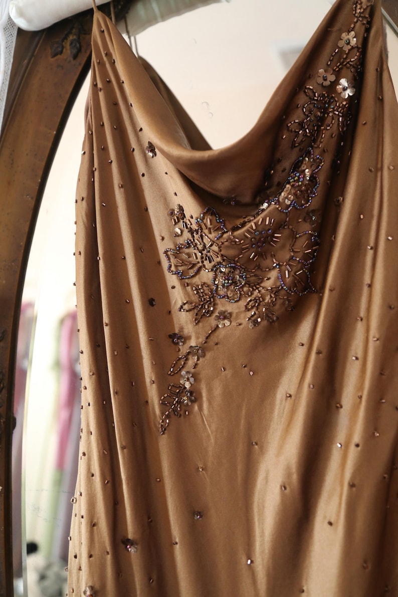 May include: A flowing, champagne-colored dress with a cowl neckline. The dress is adorned with intricate beadwork and sequins in shades of brown and silver, creating a shimmering effect. The fabric appears to be a soft, draping material.