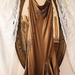 May include: A brown satin dress with a cowl neckline and a flowing, asymmetrical hemline. The dress is embellished with beaded accents and hangs on a white padded hanger in front of a vintage mirror.