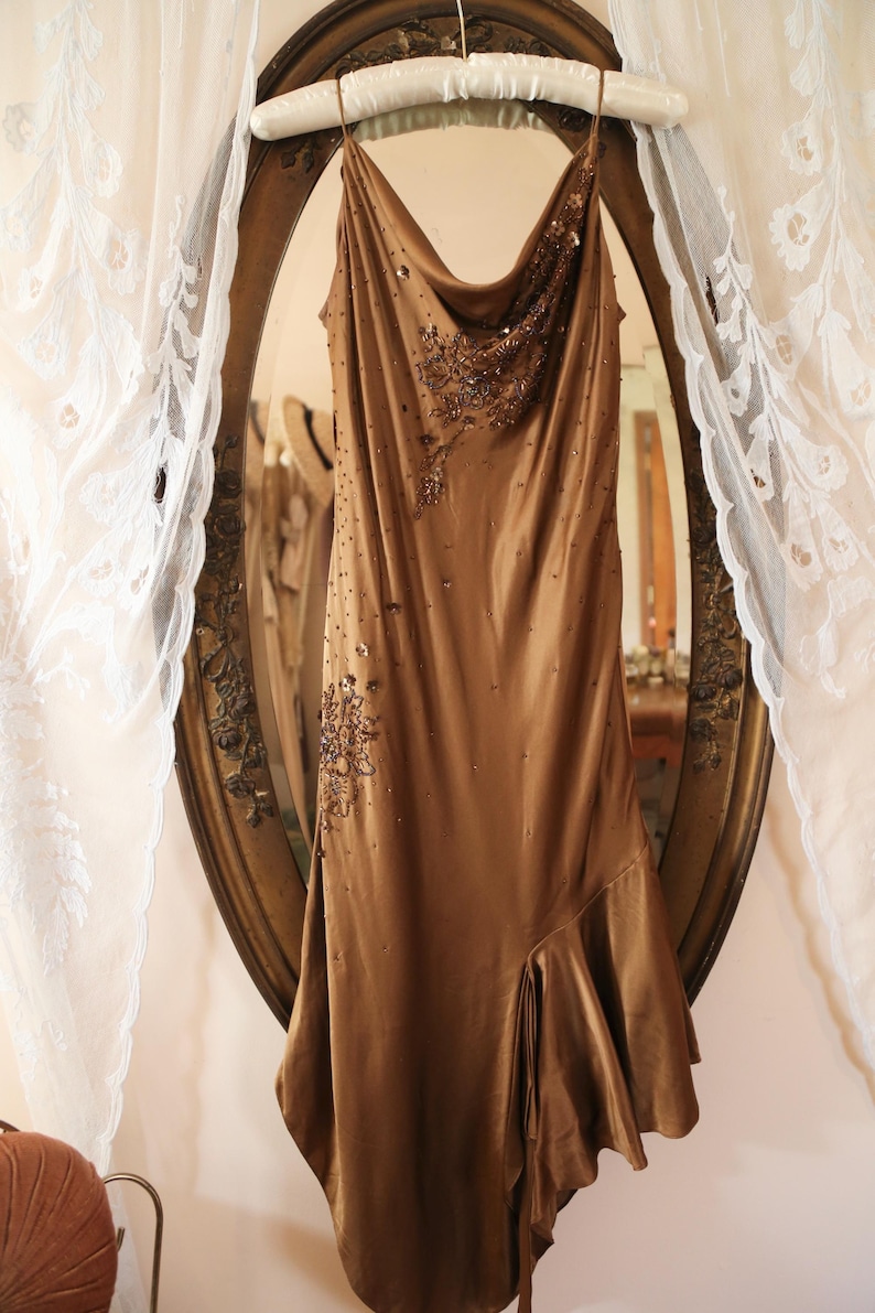 May include: A brown satin dress with a cowl neckline and a flowing, asymmetrical hemline. The dress is embellished with beaded accents and hangs on a white padded hanger in front of a vintage mirror.