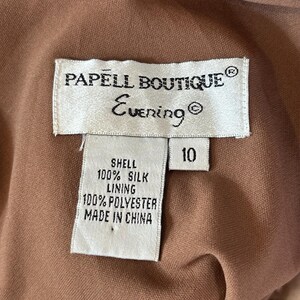 May include: Close-up of a Papell Boutique Evening dress tag. The tag reads "PAPELL BOUTIQUE Evening" and lists the shell as 100% silk and the lining as 100% polyester. The size is 10. The dress is a light brown color.