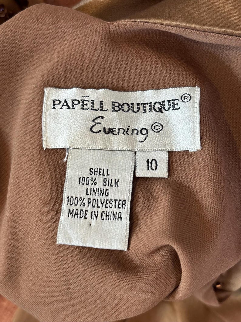 May include: Close-up of a Papell Boutique Evening dress tag. The tag reads "PAPELL BOUTIQUE Evening" and lists the shell as 100% silk and the lining as 100% polyester. The size is 10. The dress is a light brown color.