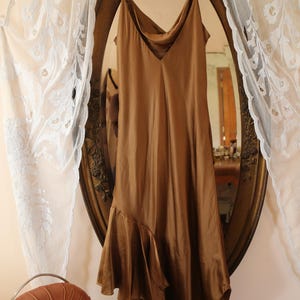 May include: A brown satin slip dress with spaghetti straps hangs in front of a vintage oval mirror. The dress has a cowl neckline and an asymmetrical hemline with a ruffled detail. The mirror is framed in a gold ornate design.