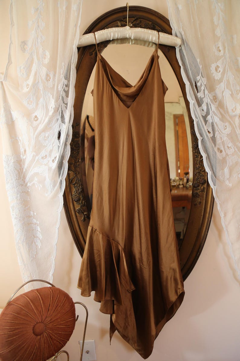 May include: A brown satin slip dress with spaghetti straps hangs in front of a vintage oval mirror. The dress has a cowl neckline and an asymmetrical hemline with a ruffled detail. The mirror is framed in a gold ornate design.