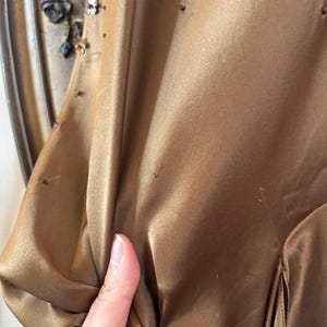 May include: A close-up of a bronze-colored satin dress. The dress has a draped design and is embellished with small, scattered beads. The sleeve is visible, and the fabric is being held by a hand. The dress appears to be vintage.