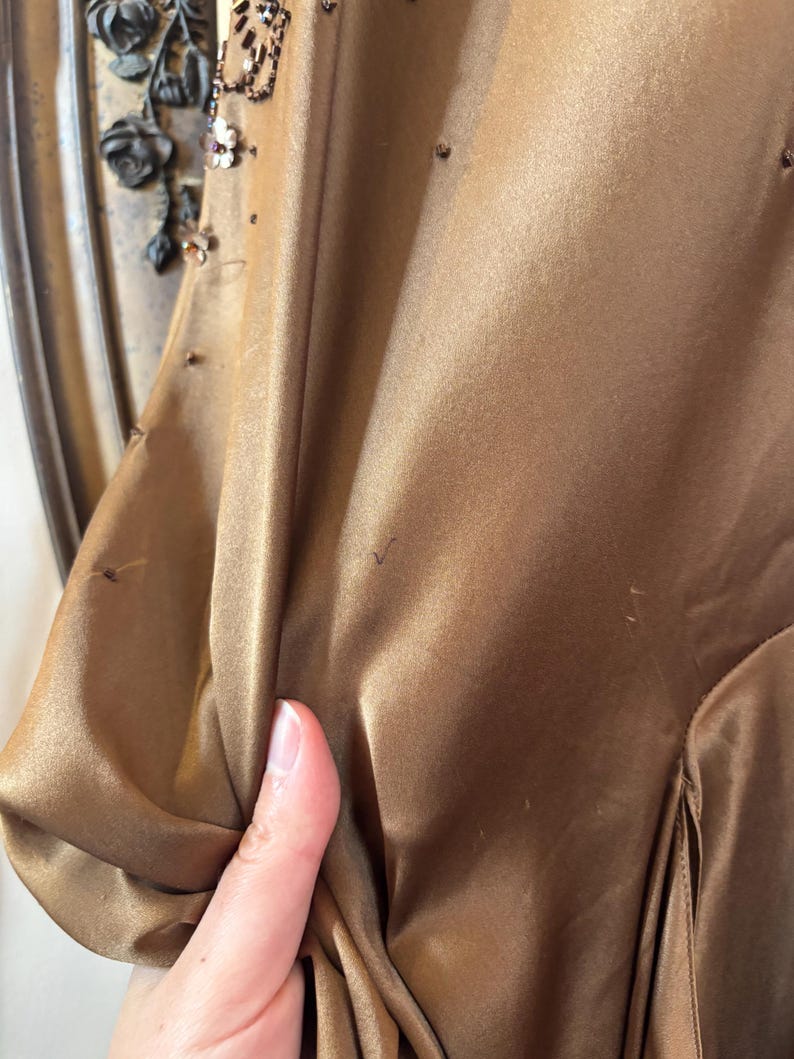 May include: A close-up of a bronze-colored satin dress. The dress has a draped design and is embellished with small, scattered beads. The sleeve is visible, and the fabric is being held by a hand. The dress appears to be vintage.