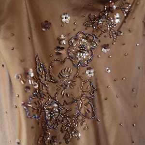 May include: A close-up of a tan-colored dress with intricate floral embroidery. The embroidery features sequins, beads, and seed beads in shades of silver, brown, and blue. Scattered sequins add sparkle to the fabric.
