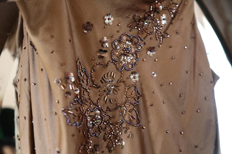 May include: A close-up of a tan-colored dress with intricate floral embroidery. The embroidery features sequins, beads, and seed beads in shades of silver, brown, and blue. Scattered sequins add sparkle to the fabric.