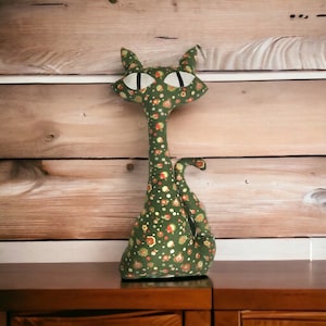 May include: A green fabric cat with a long neck and a tail curled around its body. The cat is decorated with a pattern of small green, yellow, and red circles and flowers.