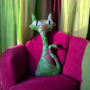 May include: A green cat-shaped plush toy with a floral pattern sits on a pink velvet armchair. The cat has large white eyes and a long neck.