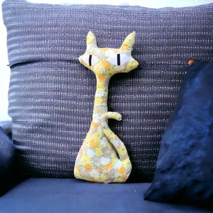 May include: A yellow and white floral patterned cat plush toy with black eyes and a white nose. The toy is sitting on a blue and white striped pillow.
