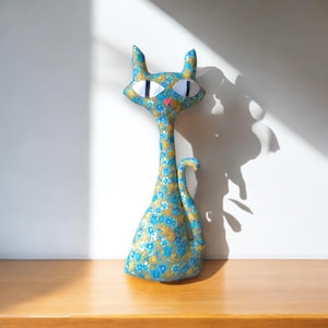 May include: A blue and gold floral patterned cat plush toy with large white eyes and a pink nose. The cat is standing on a wooden surface.