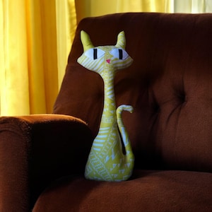 May include: A yellow and white patterned cat plush toy with large black eyes sits on a brown couch. The cat has a long neck and a long tail.