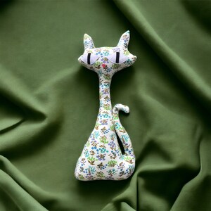 May include: A white cat plush toy with a floral pattern in shades of green and blue. The cat has large, black eyes and a pink nose.