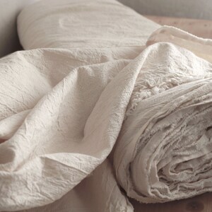 May include: A close-up of a roll of off-white fabric with a textured surface. The fabric is folded and rolled, creating a soft, rounded shape. The fabric has a frayed edge.