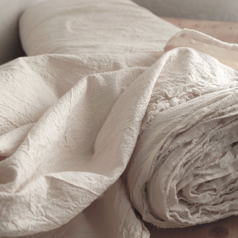 May include: A close-up of a roll of off-white fabric with a textured surface. The fabric is folded and rolled, creating a soft, fluffy appearance.