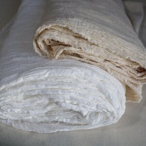 May include: Two rolls of white fabric, one is a natural off-white color and the other is a bright white. Both fabrics are folded and have a slightly rough texture.