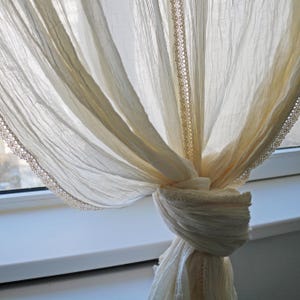 May include: Cream-colored sheer curtain with a delicate lace trim. The curtain is tied in a knot, creating a soft, draped effect. The fabric appears lightweight and airy, perfect for filtering sunlight. The lace trim adds a touch of elegance.