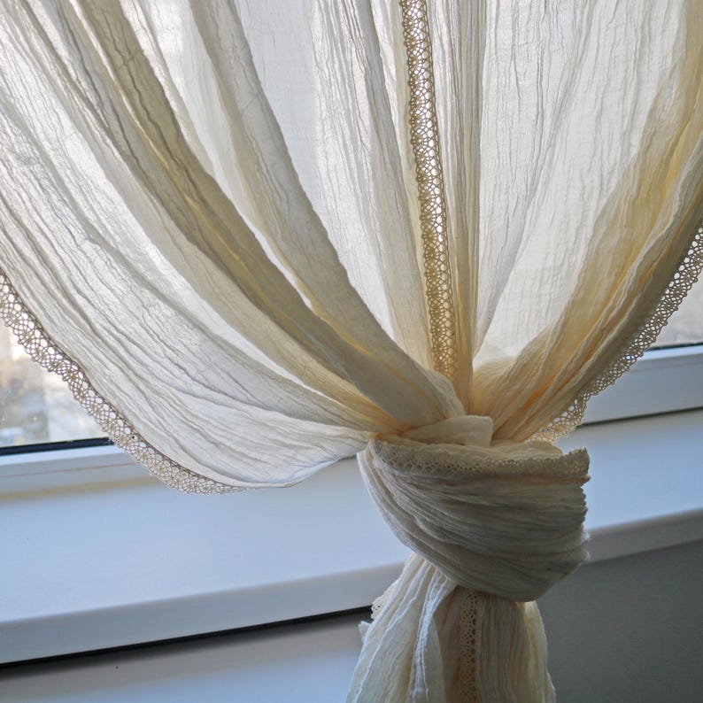 May include: Cream-colored sheer curtain with a delicate lace trim. The curtain is tied in a knot, creating a soft, draped effect. The fabric appears lightweight and airy, perfect for filtering sunlight. The lace trim adds a touch of elegance.