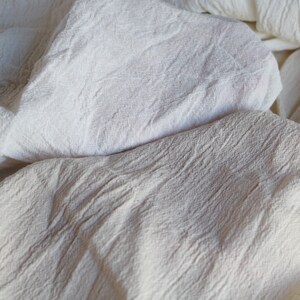 May include: A close-up of a white cotton fabric with a textured surface. The fabric is folded and wrinkled, creating a soft and natural look.