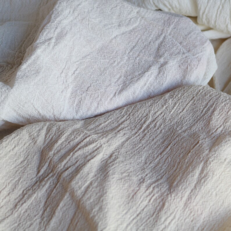 May include: A close-up of a white cotton fabric with a textured surface. The fabric is folded and wrinkled, creating a soft and natural look.