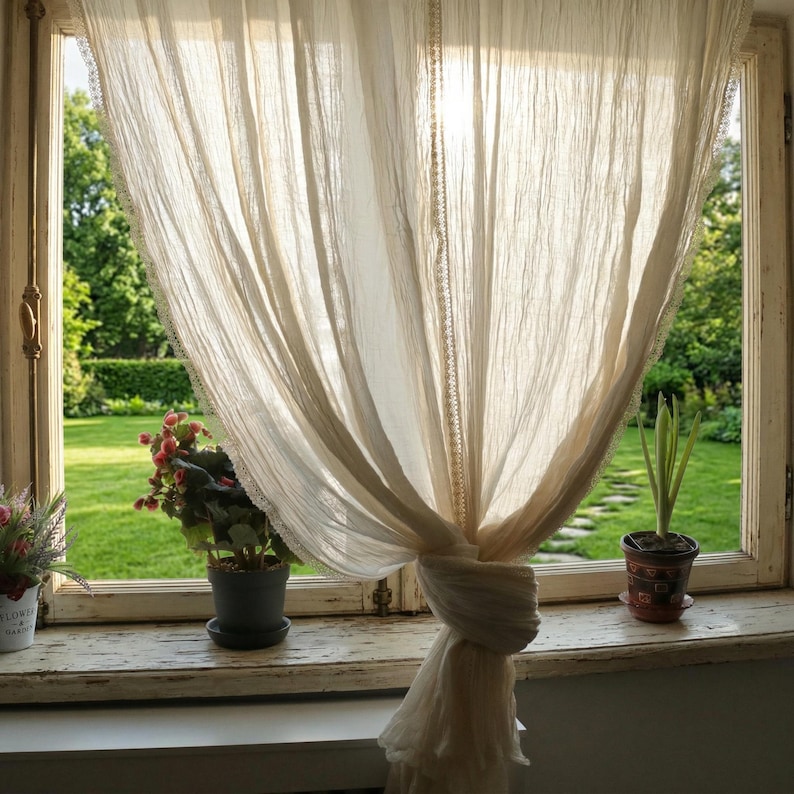 May include: Sheer, cream-colored curtain tied back, framing a sunlit garden view. The curtain has a delicate lace trim. A weathered windowsill holds potted plants with red and green foliage, adding a touch of nature to the scene. The window is framed by a white, distressed wooden frame.