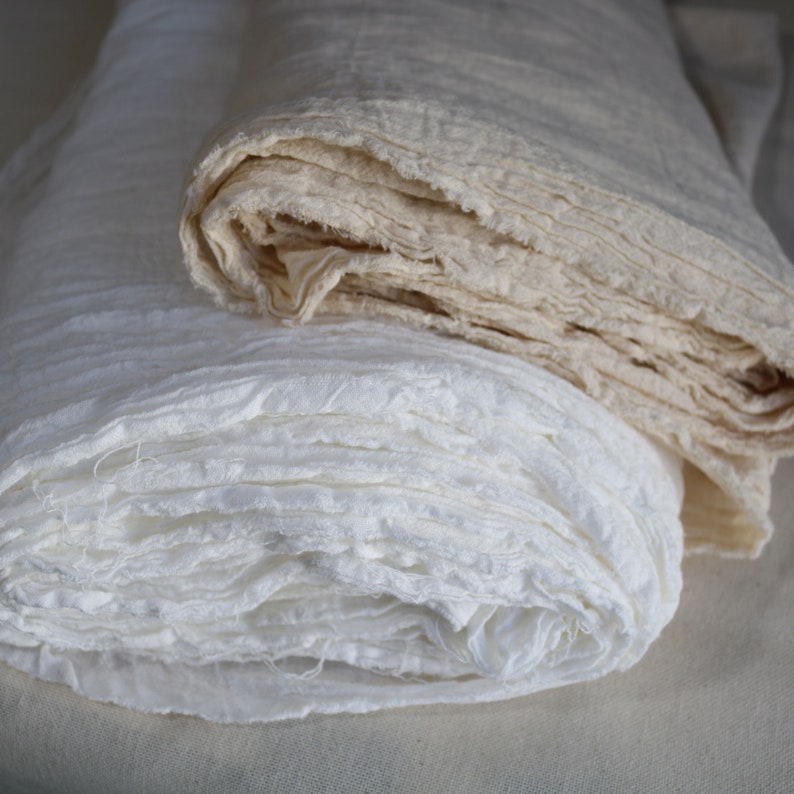 Puede incluir: Dos rollos de tela blanca, uno es de color blanco roto natural y el otro es blanco brillante. Ambas telas est&aacute;n dobladas y tienen una textura ligeramente rugosa.