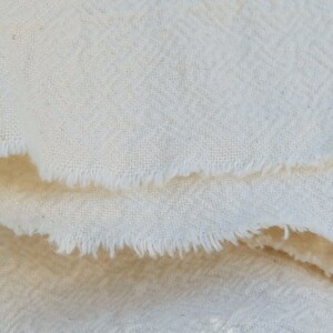 May include: Close-up of a white fabric with a textured weave. The fabric has a raw edge and is folded over.