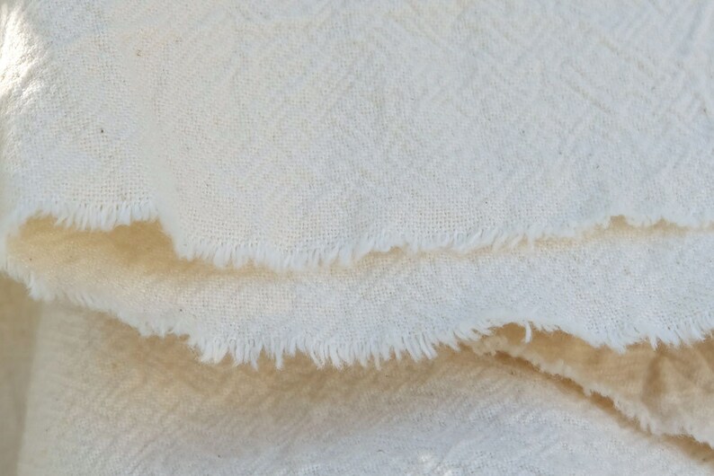 May include: Close-up of a white fabric with a textured weave. The fabric has a raw edge and is folded over.