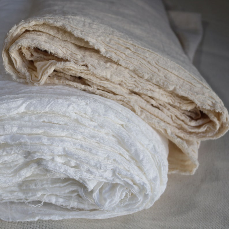 May include: Two rolls of white fabric, one is a lighter beige color and the other is a bright white. Both are made of a soft, gauzy material and are rolled up tightly.