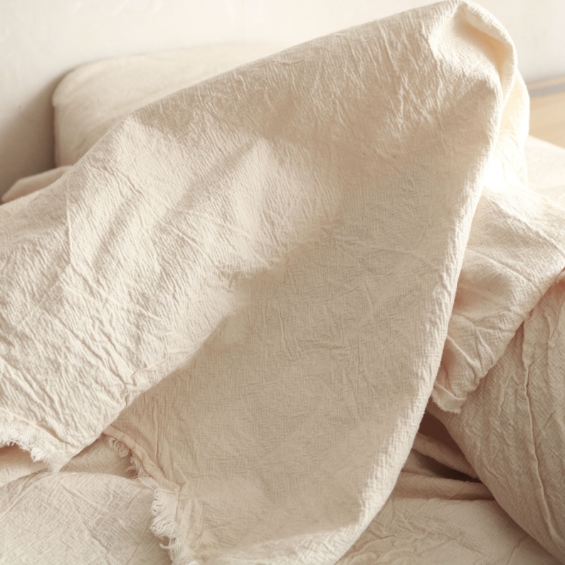 May include: A white cotton throw blanket with a textured, crinkled surface. The blanket is folded and draped over a bed.