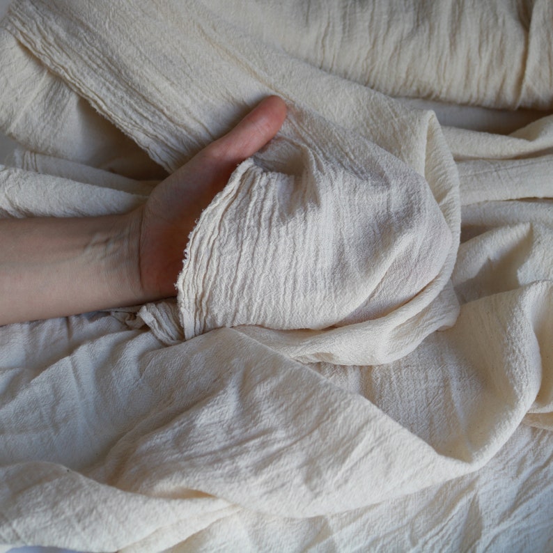 May include: A close-up of a hand holding a piece of off-white fabric. The fabric is soft and wrinkled, with a textured surface.