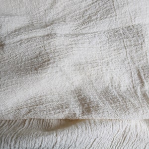 Puede incluir: Primer plano de una tela blanca texturizada con un aspecto suave y arrugado. La tela est&aacute; doblada por la mitad, creando una superficie lisa y uniforme.